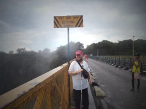 A man stands on a bridge with a sign overhead reading 'You are now entering Zambia.' He wears a red cap, white shirt, and is holding a walking cane. The bridge is wet, and mist can be seen in the background, likely from the nearby Victoria Falls. Another person stands further down the bridge taking a photo.