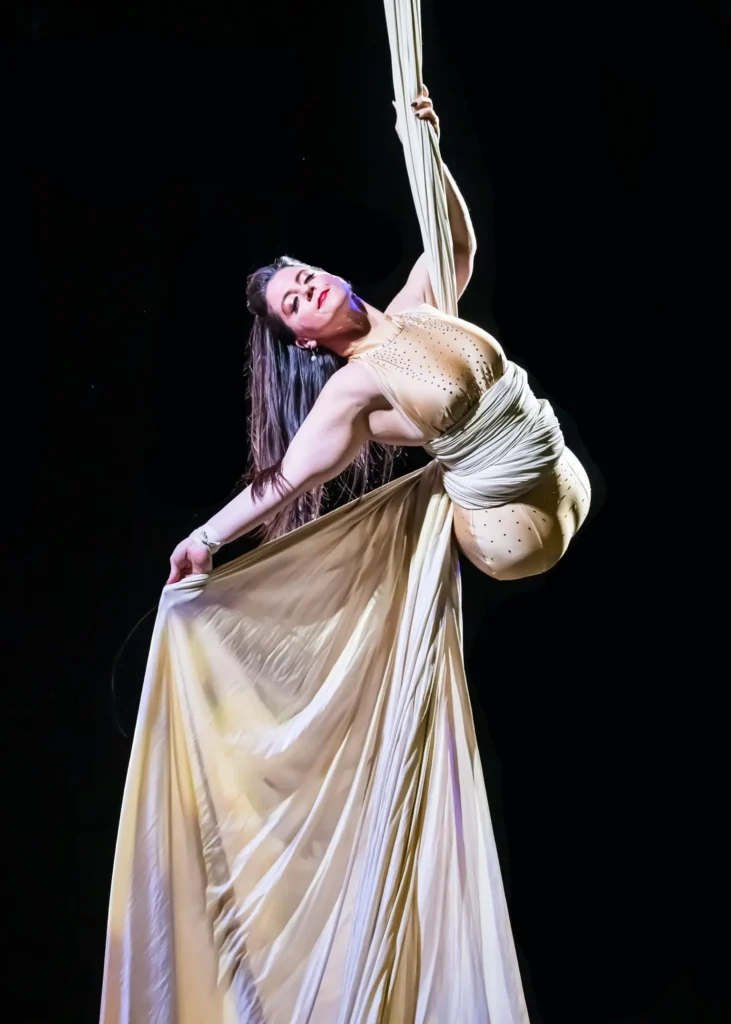 A woman with no logs, performing an aerial silk routine against a black background. She is suspended mid-air, holding onto white silk fabric, which wraps around her torso and flows outward. She wears a fitted, sleeveless costume adorned with subtle embellishments. Her head is tilted back gracefully, with long hair flowing behind, and one arm extended, adding elegance to her pose.