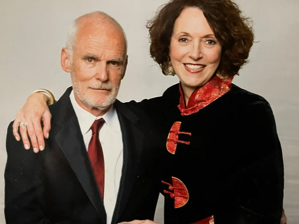 This is a portrait of an older couple. The man, with short, white hair and a beard, is wearing a formal black suit with a white shirt and a red tie. The woman next to him has curly, shoulder-length brown hair and is smiling. She is dressed in a black outfit with striking red and gold accents, which resembles traditional Chinese or Asian-inspired clothing. The woman has her arm around the man, and they are standing closely together, both facing the camera.