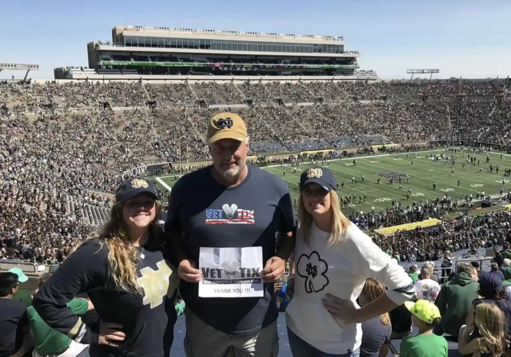 A group of three people is standing in a football stadium, with the field and crowd visible in the background. The person in the middle, wearing a navy blue Vet Tix shirt and a Notre Dame cap, is holding a sign that says 'Vet Tix' with a thank you message. The two people on either side, both wearing Notre Dame apparel, are smiling and posing with their hands on their hips. The stadium is filled with spectators, and the atmosphere is lively, indicating it is a game day.