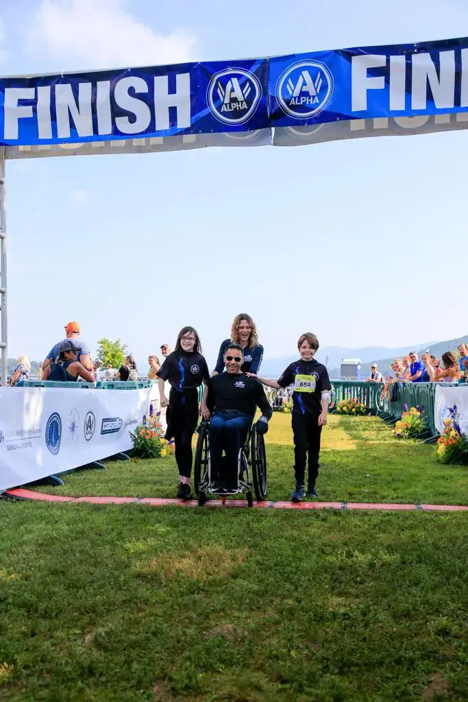 The image shows a man in a wheelchair crossing the finish line of a race, accompanied by three other people: two children standing on either side of him and a woman behind him. The finish line banner above them displays the word "FINISH" and logos with the word "ALPHA" on either side. The people in the image appear to be smiling, and there are spectators and greenery in the background. It seems to be a positive moment at an outdoor event or race, with the participants celebrating their achievement together.