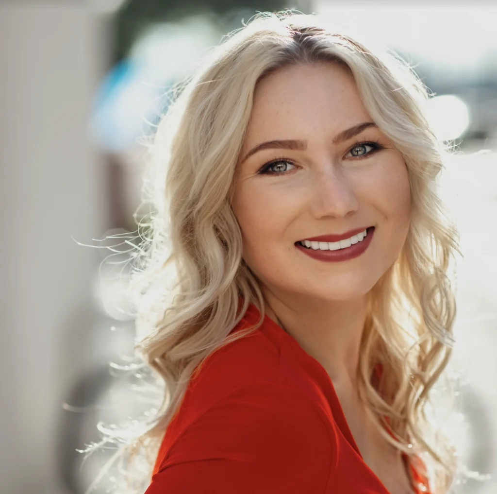 A blonde woman with loose, wavy hair smiles brightly at the camera. She is wearing a vibrant red top, and the sunlight enhances her features, creating a warm, inviting atmosphere. The background is softly blurred, bringing focus to her cheerful expression and sparkling eyes.