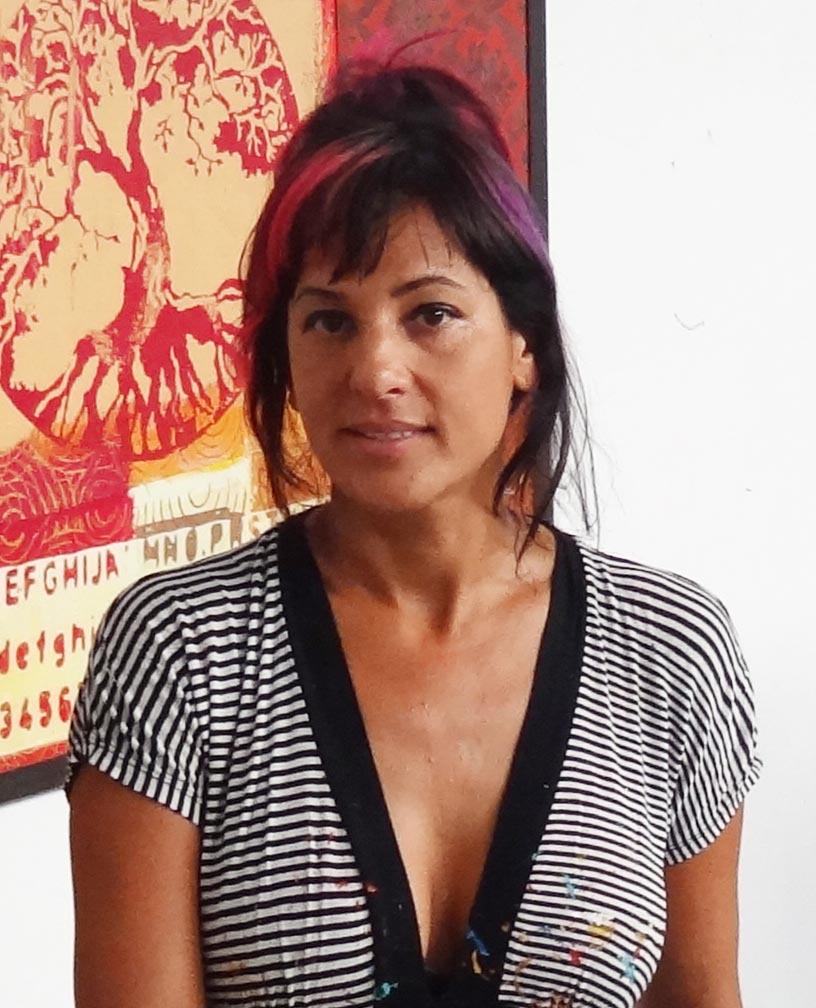 Elisa Lejuez, a contemporary artist, standing in front of one of her vibrant, patterned artworks. She has dark hair with streaks of purple and pink, wearing a striped black and white top with paint splatters. The background art piece features bold, abstract designs in red and orange tones.