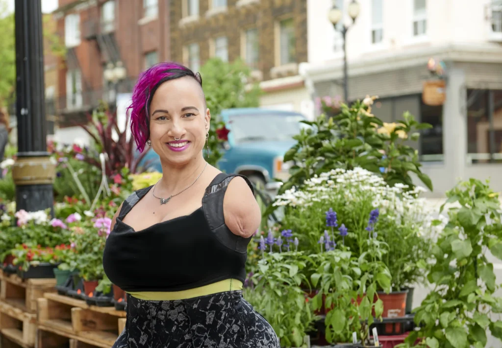 A confident woman with a shaved head and vibrant pink hair smiles warmly in an outdoor setting with various colorful flowers and plants in the background. She wears a black dress with a patterned skirt and displays a sense of style and empowerment.