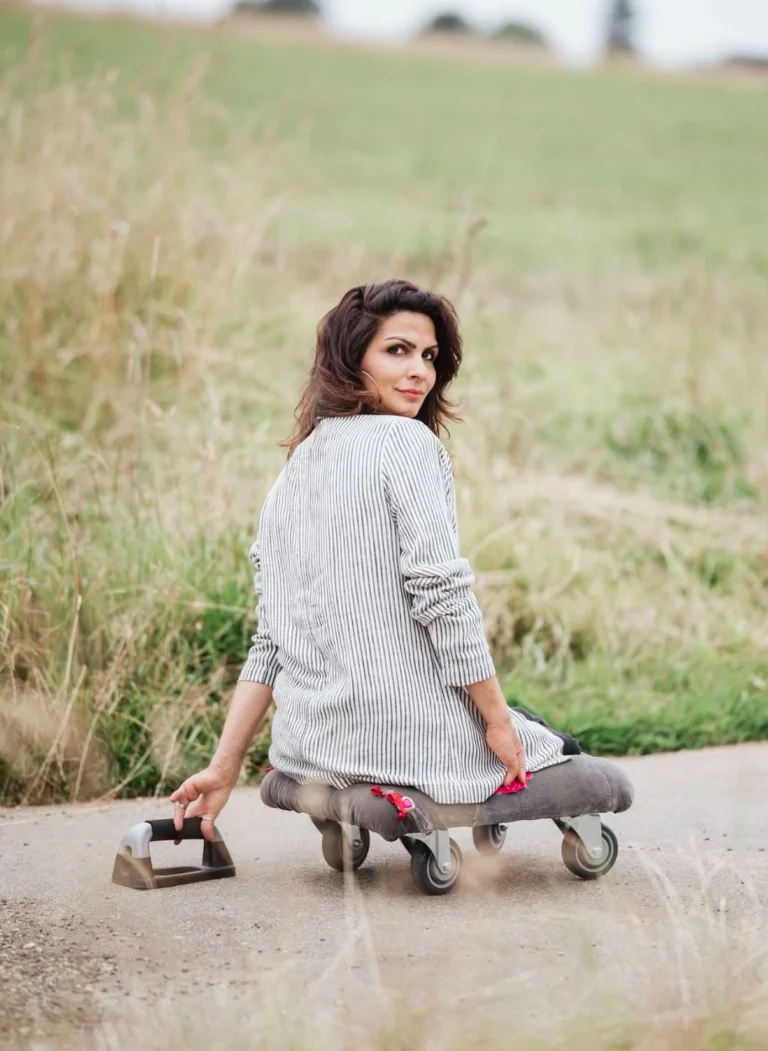 A woman sitting on a wheeled board on a paved path, holding a handle for support, surrounded by a grassy field. She is looking back over her shoulder with a slight smile, wearing a striped shirt, and appears relaxed in the natural outdoor setting.