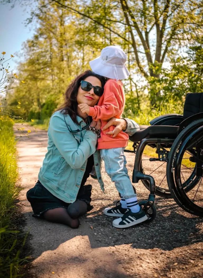 A mother wearing sunglasses and a denim jacket kneeling on a sunny path while hugging her young child dressed in a coral sweatshirt, light blue jeans, and a white sun hat. The child is standing next to a wheelchair, surrounded by greenery and wildflowers, under a bright and warm natural light.