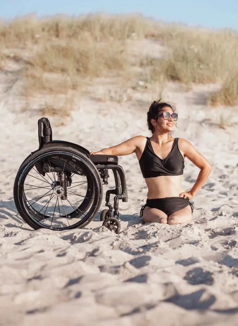 Person sitting on the sandy beach, wearing a black swimsuit and sunglasses, smiling confidently with a wheelchair beside them, with dunes and a clear blue sky in the background