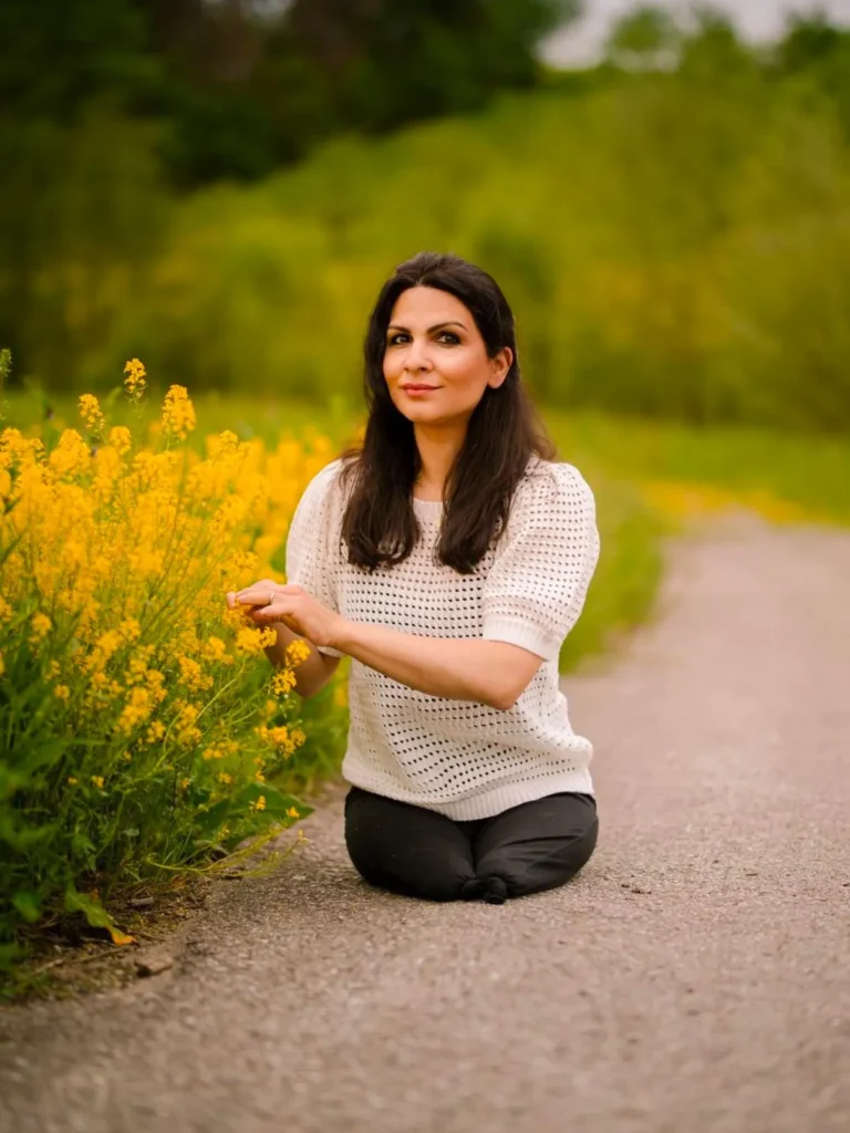 A woman with long dark hair sits on her knees on a paved pathway surrounded by greenery and vibrant yellow flowers. She is wearing a white short-sleeve sweater and black pants, smiling softly while gently touching the flowers. The background is lush and blurred, emphasizing the natural setting.