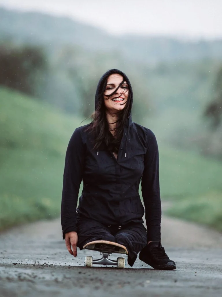 Person with long dark hair smiling and wearing a black outfit while sitting on a skateboard in a serene outdoor setting with green hills and a soft, overcast sky in the background.