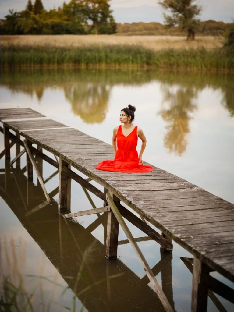 Person in a bright red dress sitting on a rustic wooden dock over a calm lake, surrounded by lush greenery and trees, with reflections visible on the water