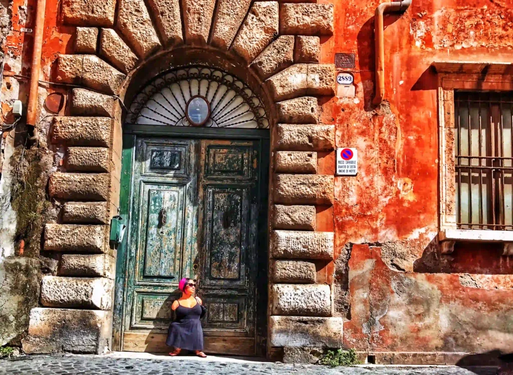 An artistic photograph of a weathered green wooden door set in an arched stone frame, with rich textures of worn brick and orange plaster surrounding it. A person wearing a dark dress and pink headscarf sits casually on the steps in front of the door, bathed in warm sunlight. The scene exudes a rustic charm, with vibrant colors and a touch of urban decay