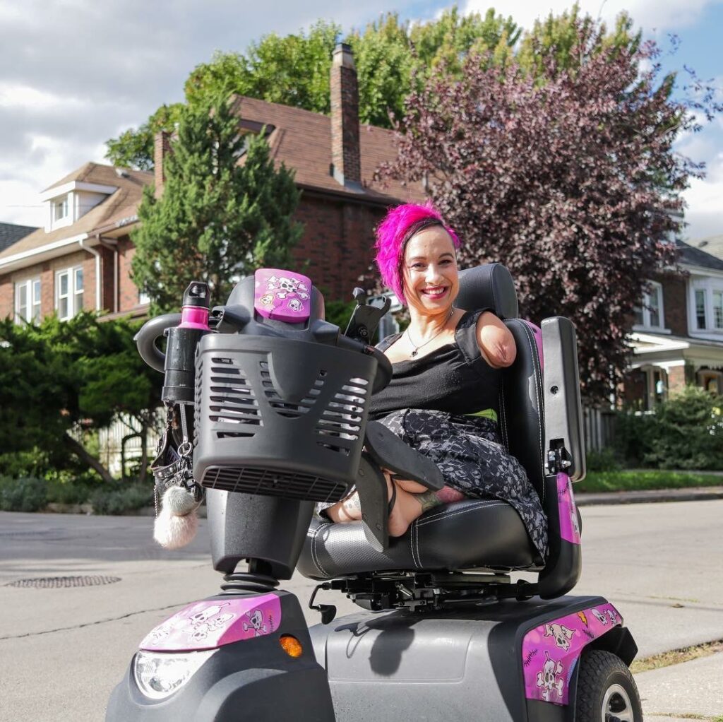 A smiling person with bright pink hair sits on a mobility scooter adorned with playful pink designs and decals. They are outdoors on a sunny day, with residential houses and trees in the background. The person has a confident and cheerful demeanor, wearing a black outfit and a patterned skirt.