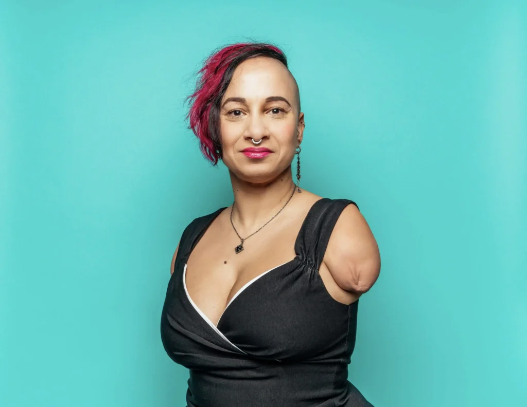 Portrait of a confident woman with a teal background, featuring a stylish asymmetrical hairstyle with pink highlights, a septum piercing, and a black sleeveless dress. She is smiling warmly, embracing her unique beauty and proudly showing her armless physique.