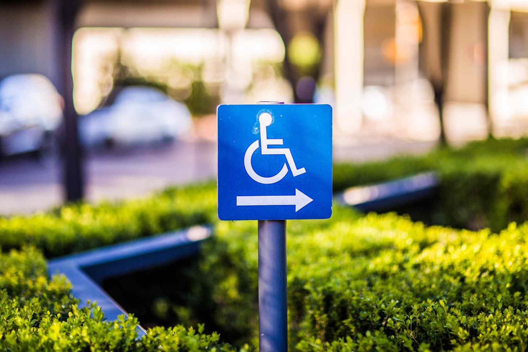 Blue accessibility sign with a white wheelchair symbol and an arrow pointing to the right, placed among green shrubs with a blurred urban background.