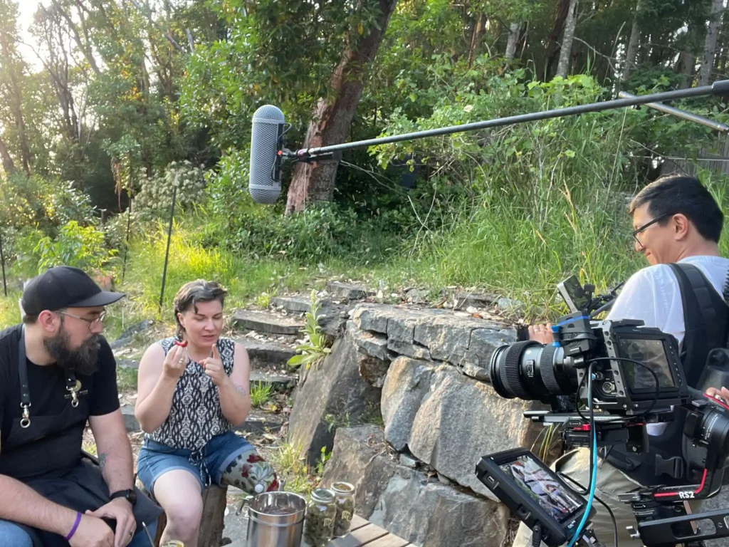 Outdoor filming setup featuring two individuals seated on stone steps surrounded by greenery. A boom microphone is positioned overhead, and a camera crew operates professional video equipment, capturing the scene.