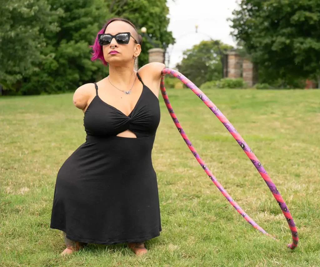 A confident individual with short, vibrant pink and black hair stands outdoors in a grassy field. She is wearing a black dress and sunglasses while holding a colorful pink and purple hula hoop, radiating empowerment and joy.