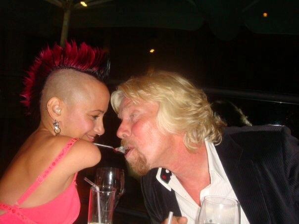 Two individuals sharing a moment of fun at a social event. The person on the left has a bright red mohawk hairstyle, shaved sides, and is wearing a pink outfit, while the person on the right has blonde hair, a beard, and is wearing a suit jacket. They appear to be playfully interacting, with a spoon involved.