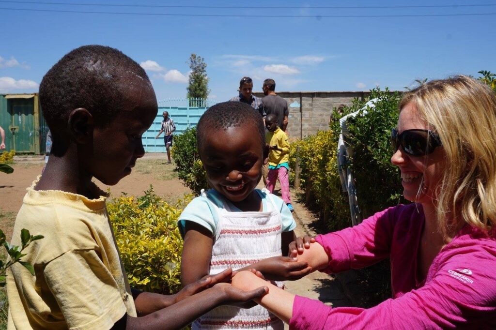 Two children and an adult woman smiling and playing together outdoors on a sunny day, with bushes, a gate, and other people visible in the background."