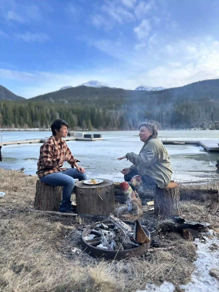 Two people are sitting on tree stump seats beside a campfire, engaged in conversation. They are outdoors near a frozen lake surrounded by dense evergreen forest and snow-capped mountains. The sky is clear and blue, adding to the serene, natural setting. A plate of food rests on a tree stump table between them, and smoke rises gently from the fire pit in the foreground.