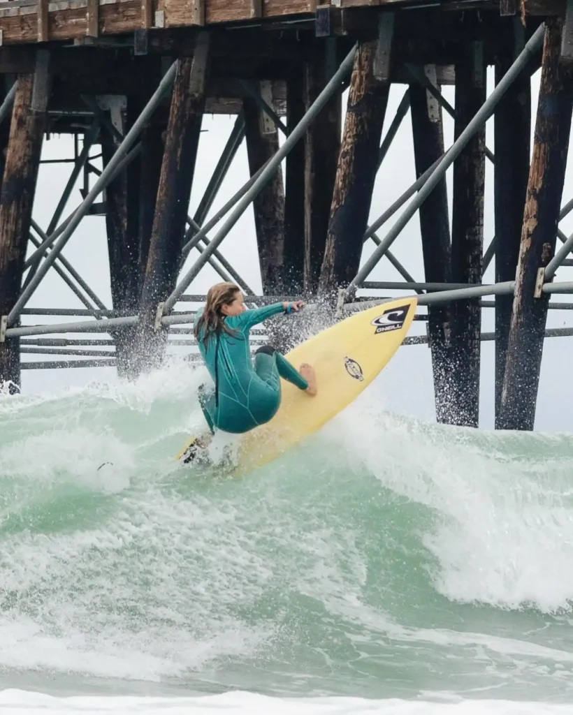 A surfer wearing a teal wetsuit rides a yellow surfboard on a wave near a wooden pier. The surfboard is tilted upward as the surfer maneuvers through the choppy water, with splashes and foam surrounding the scene. The wooden beams of the pier provide a dramatic backdrop.