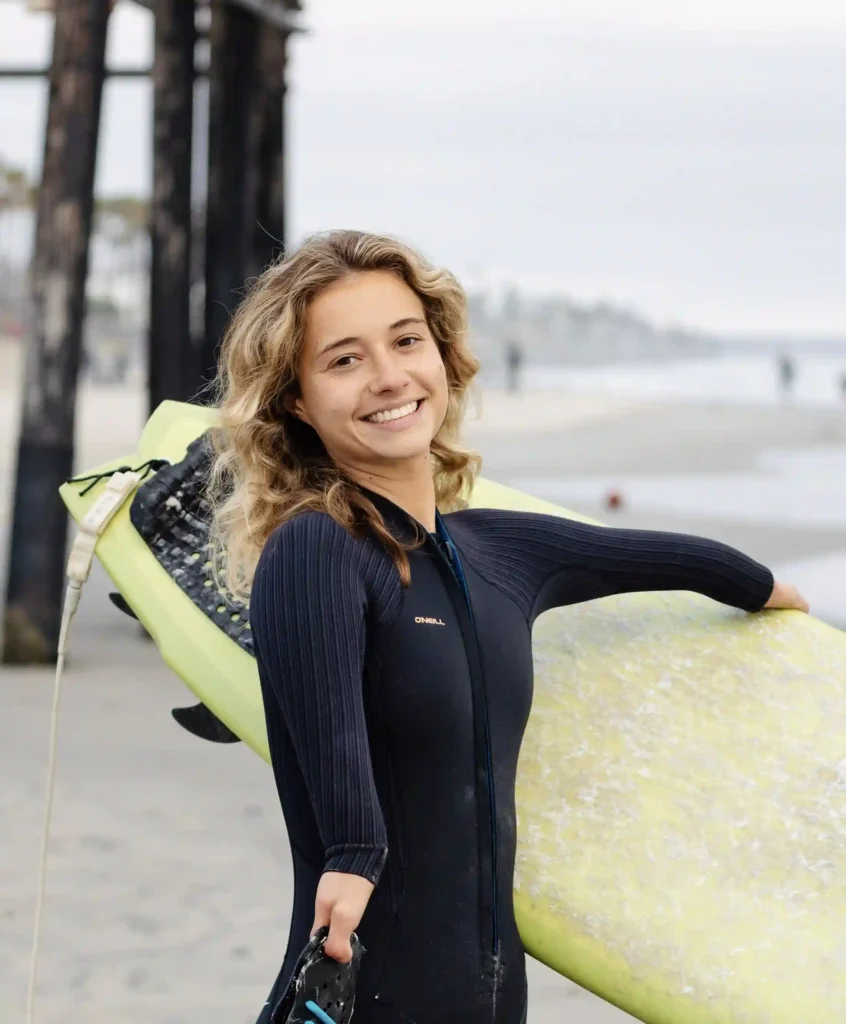 Young woman with curly blonde hair smiling and holding a yellow surfboard under her arm. She is wearing a black wetsuit and standing on a beach with the ocean and a pier visible in the background.