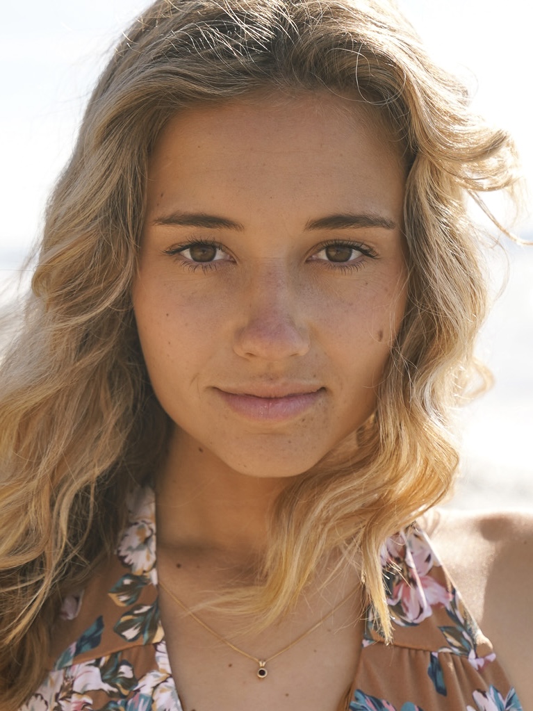 A portrait of a young woman with wavy, light brown hair and natural makeup, wearing a floral-patterned top and a delicate gold necklace, looking directly at the camera with a calm expression. The background appears softly lit, suggesting an outdoor setting with bright sunlight.