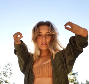 A young woman with blonde, wavy hair is gazing into the camera under a clear blue sky. She is wearing a textured olive green shirt and a rust-colored ribbed crop top. Her hands are raised near her face, creating a dynamic pose. Sunlight illuminates her hair and face, giving the image a warm and natural feel. In the background, there are faint trees out of focus.