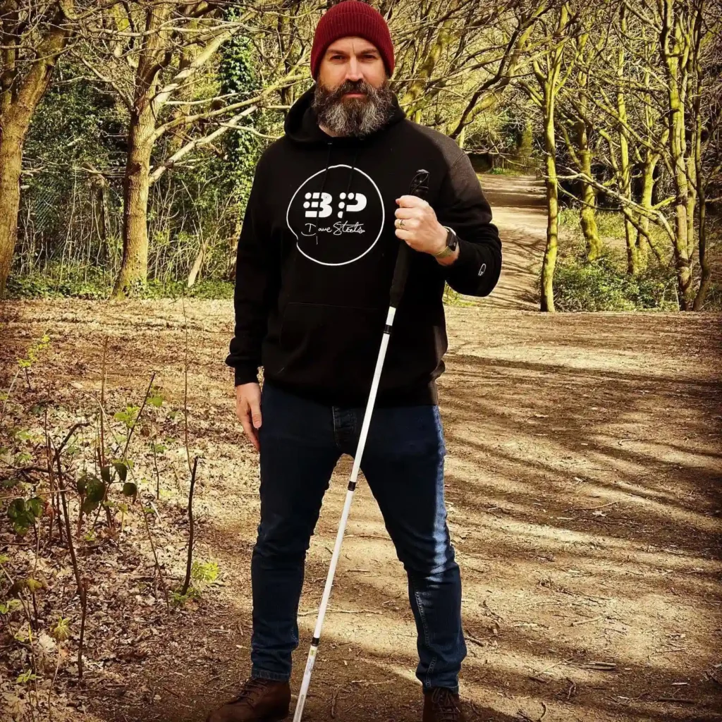 Man with a beard wearing a beanie, dark hoodie, and jeans, holding a white cane while standing on a wooded trail surrounded by trees