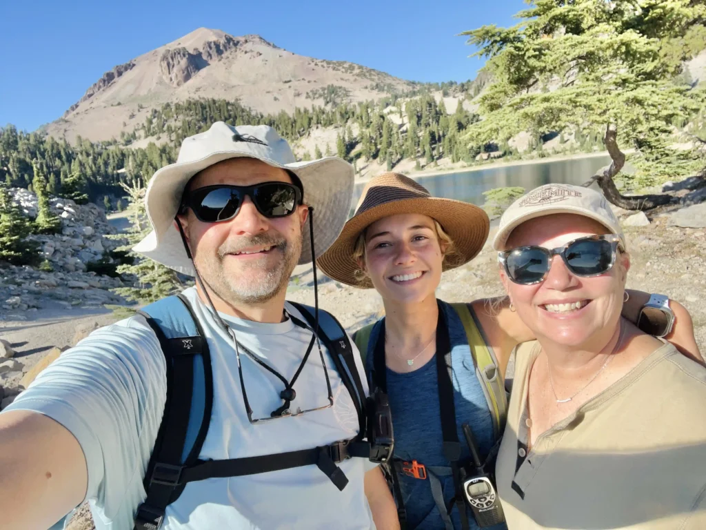 A group photo of three hikers posing happily outdoors in front of a scenic mountain and lake backdrop. The man on the left wears a light-colored wide-brimmed hat, sunglasses, and a backpack. The woman in the middle has a straw hat and a bright smile, also wearing a backpack. The woman on the right wears sunglasses, a beige cap, and a tan shirt, smiling warmly. The background includes evergreen trees, a calm lake, and a rugged mountain under a clear blue sky.