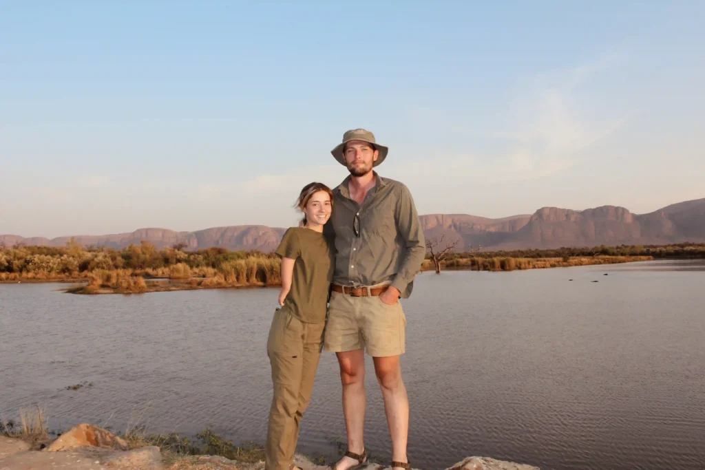 A couple posing near a calm body of water during sunset, with rugged mountains and grassy vegetation in the background. The man is wearing a wide-brimmed hat, long-sleeved shirt, and khaki shorts, while the woman is dressed in a khaki-green t-shirt and pants, both smiling warmly. The scene has a serene and adventurous safari vibe.