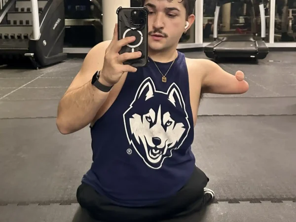 Tim Mason A man in a gym takes a selfie in front of a mirror. He has short, curly hair, a mustache, and is wearing a navy blue tank top with a husky logo. One of his arms ends above the elbow. In the background, there are treadmills and large windows, showcasing a nighttime urban view.