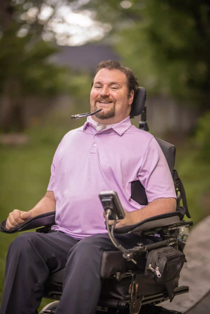 A close-up of a man in a power wheelchair, wearing a light pink polo shirt, smiling warmly. He has a breathing tube and controls the wheelchair with a mouthpiece. The background is blurred with greenery, highlighting a peaceful outdoor setting.