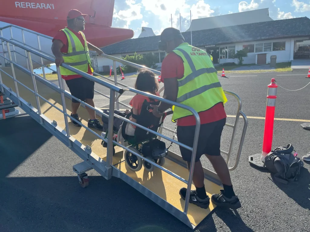 Two airport staff members assist a person in a wheelchair as they ascend a ramp towards a red aircraft under a clear blue sky.