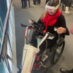 A woman in a wheelchair wears a VR headset and holds controllers, engaged in an interactive experience at a busy event.