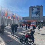 A busy outdoor scene at Messe Berlin, adorned with colorful flags, showcasing attendees mingling under a clear blue sky.