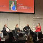 A panel discussion featuring four speakers, with a PowerPoint presentation displaying participants' names and titles in a conference setting.