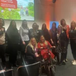 A group of women poses together, smiling, in a conference hall during a networking break. A bike is in the foreground.