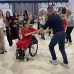 A lively group of people dance around a woman in a red dress using a wheelchair, showcasing joy and inclusivity at an event.