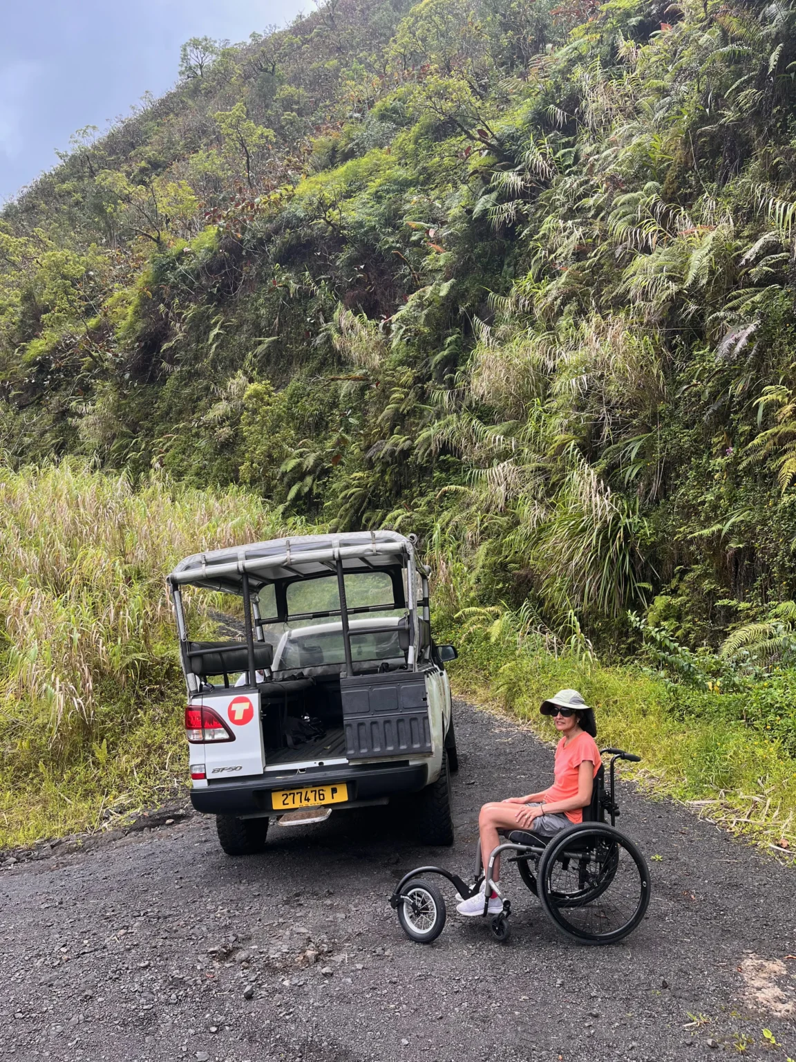 A person in a wheelchair sits on a gravel path beside an open utility vehicle, surrounded by lush green hillside vegetation.