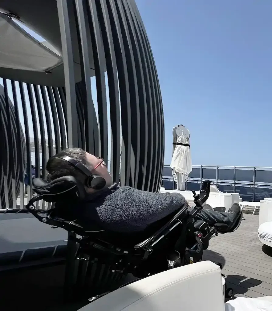 A person in a wheelchair relaxes outdoors with headphones on, against a backdrop of clear blue sky and modern architecture.