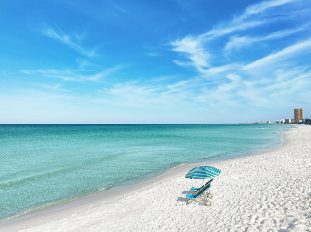 A serene beach scene featuring a blue-striped umbrella and lounge chair on white sand, with calm turquoise waters and a clear sky.