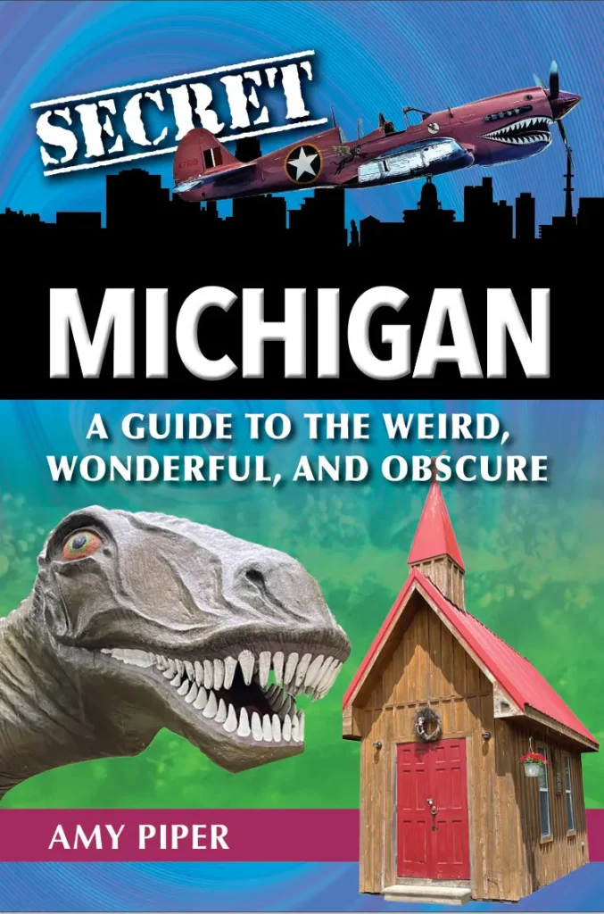 The cover of the book "Secret Michigan: A Guide to the Weird, Wonderful, and Obscure" by Amy Piper features a bold design. The top portion displays a vintage warplane with a shark mouth painted on its nose, flying against a blue sky with a black silhouette of a city skyline below. The word "SECRET" is stamped in white, distressed text. The middle of the cover prominently showcases the title "MICHIGAN" in large, white, bold letters, followed by the subtitle "A Guide to the Weird, Wonderful, and Obscure." The lower portion features an oversized dinosaur head with sharp teeth and a wide-eyed expression, alongside a small rustic wooden chapel with a steep red roof and a wreath on the red door. The background transitions from blue to green, creating a visually striking and intriguing cover.