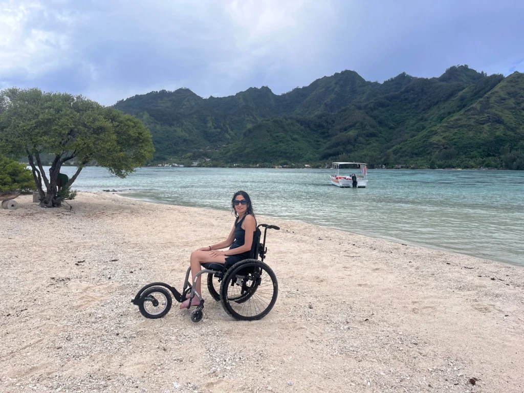 A person in a wheelchair sits on a sandy beach, with mountains and a boat in the background, under a partly cloudy sky.