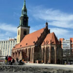 A historic church with a tall green spire, featuring red roofs and surrounded by modern buildings under a clear blue sky.