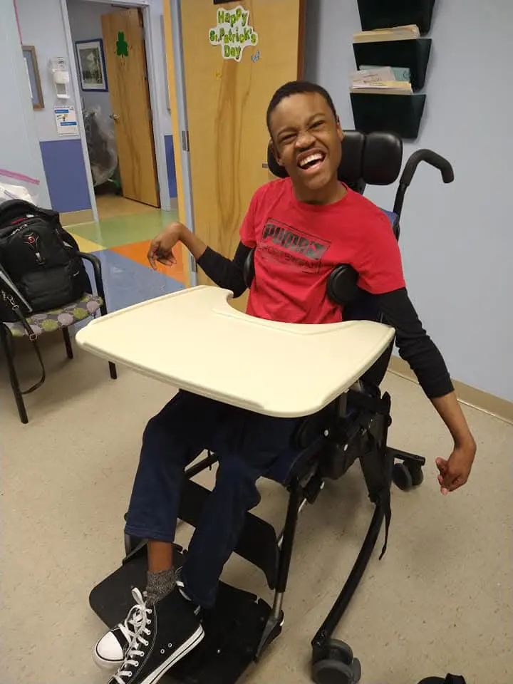 A child sits in a specialized chair with a tray, wearing a red shirt and black sneakers, in a colorful room decorated for St. Patrick's Day.