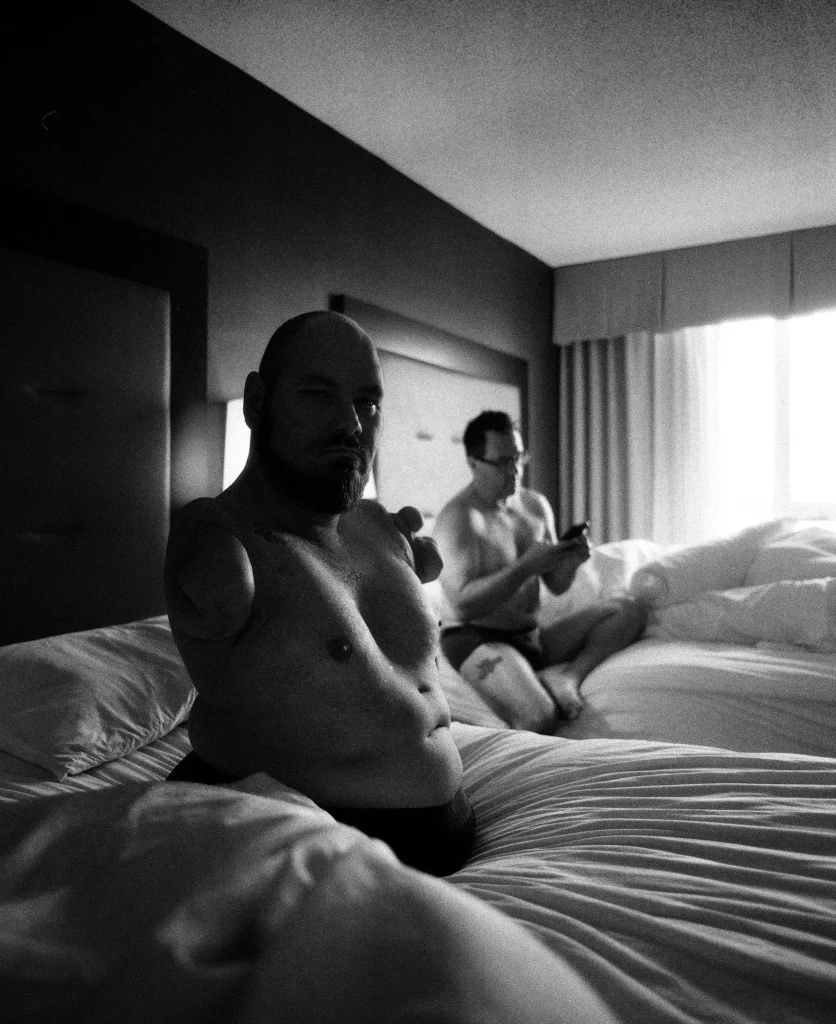 A man sitting on a bed with another man in the background, set in a bedroom with black and white decor. The scene includes bedding and a wall visible behind them.