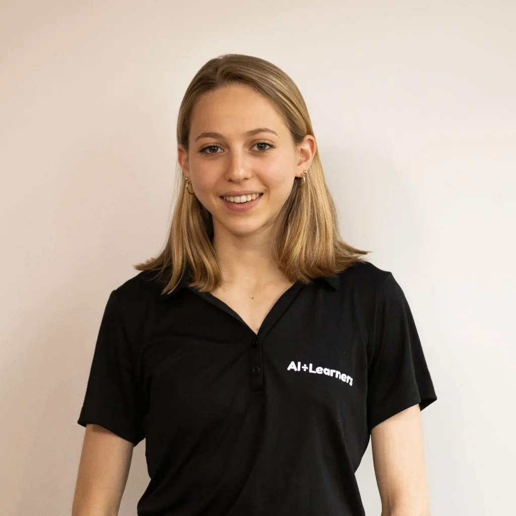 A person with shoulder-length hair wearing a black polo shirt featuring the text "AI+Learners" stands against a light-colored wall.