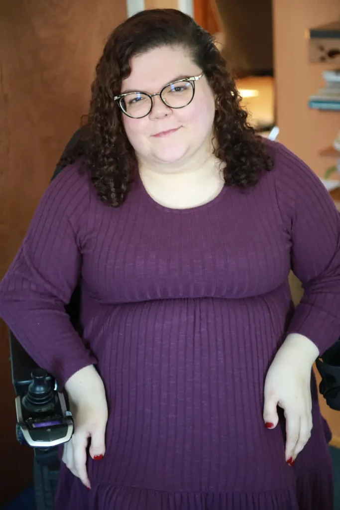 A woman in a textured purple dress sits in a wheelchair, resting her hands on her lap, with a cozy interior in the background.