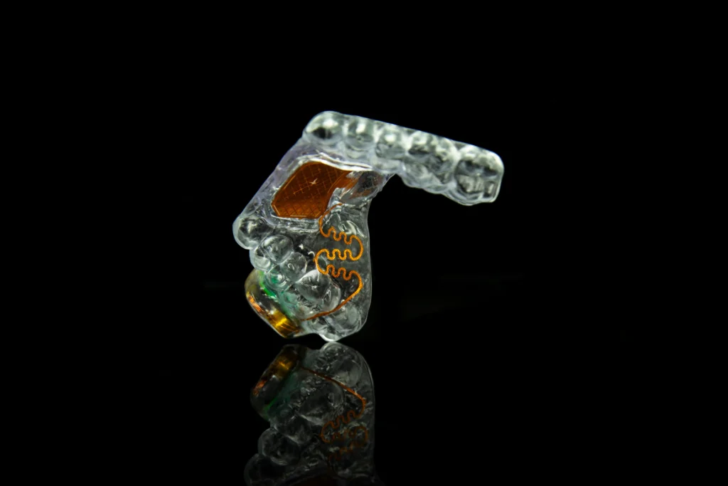 A clear, custom-fitted mouthguard with intricate electronic components, displayed on a reflective surface against a black background.