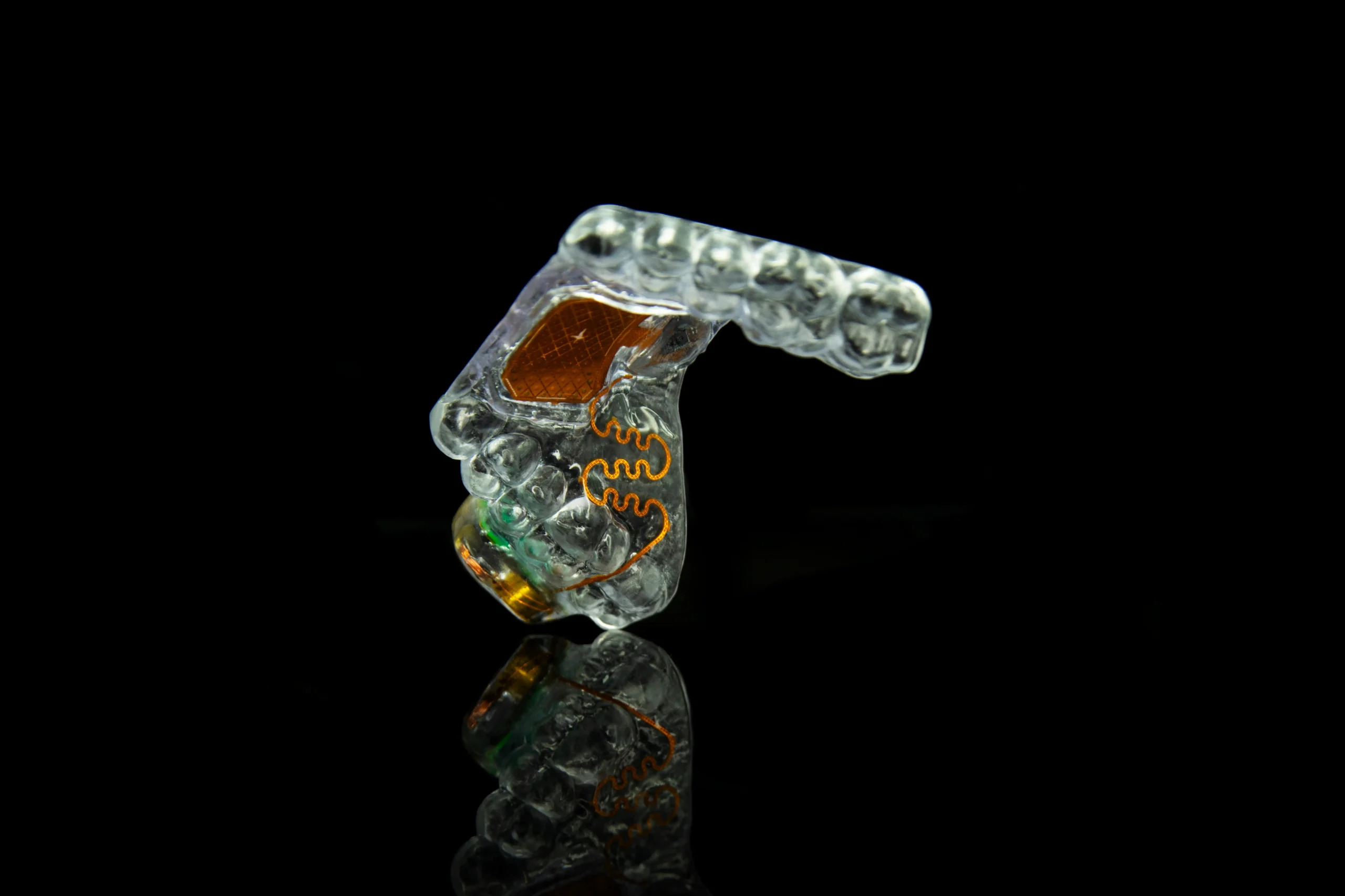 A clear, custom-fitted mouthguard with intricate electronic components, displayed on a reflective surface against a black background.
